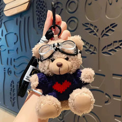 Aviator bear plush keychain wearing navy sweater with red heart and goggles, hanging from a hand