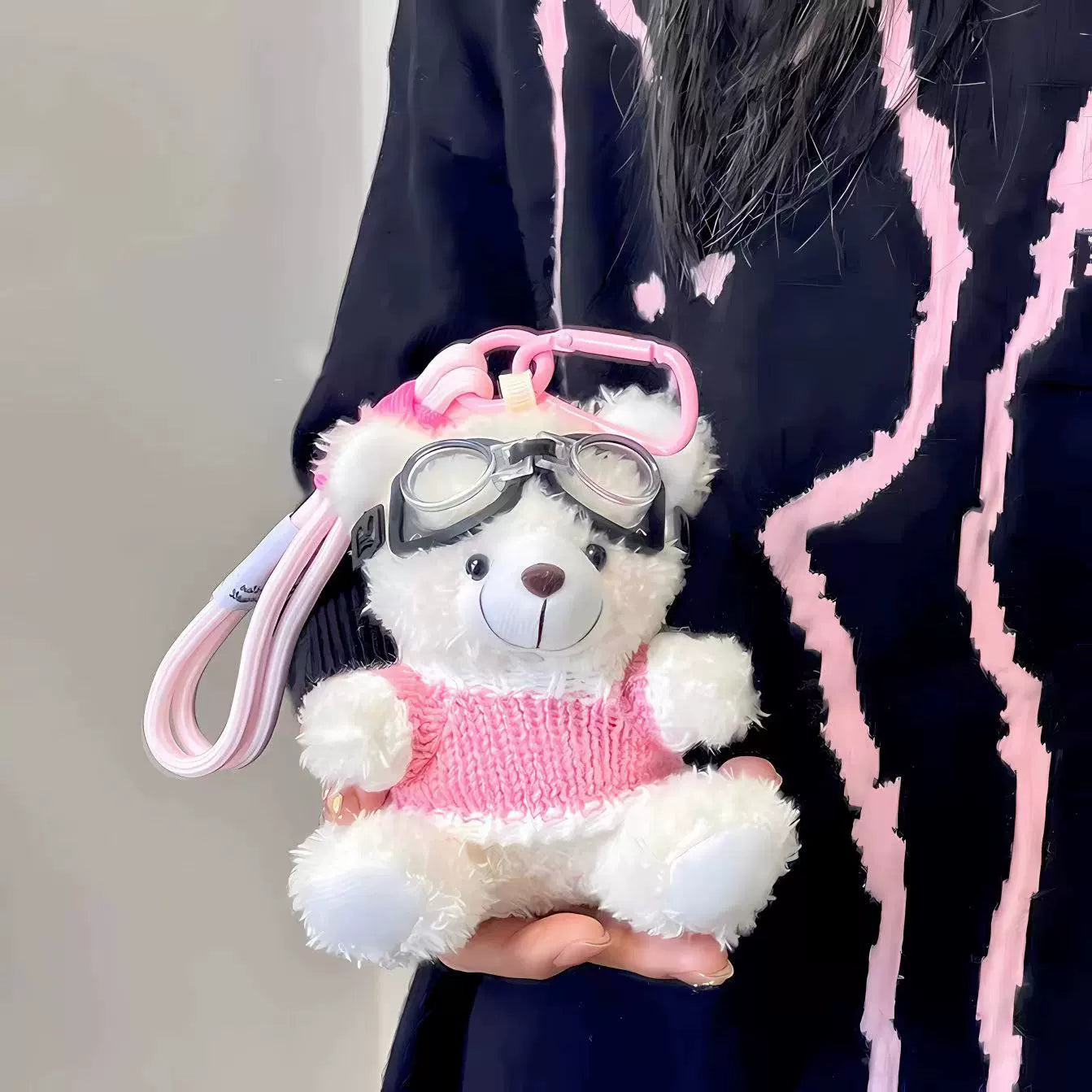 White aviator bear plush keychain in pink sweater with goggles, held close against black and pink background