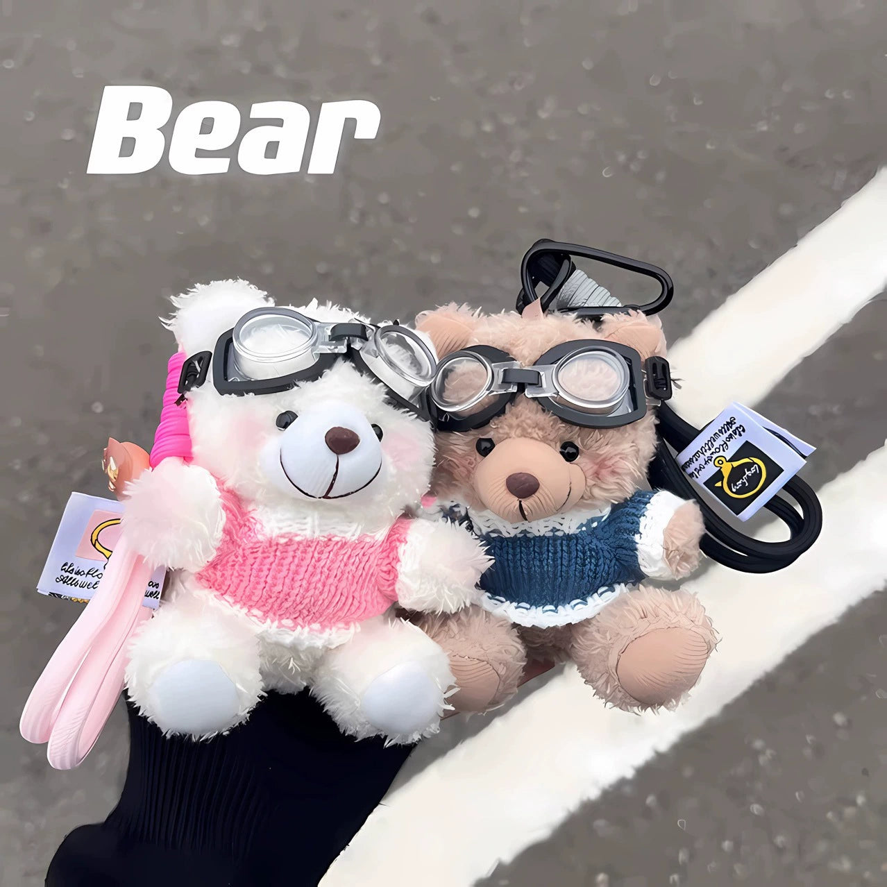 Pink and blue dressed aviator bear plush keychains, side by side, held outdoors with street background