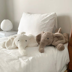 Two elephant plush toys, one white and one brown, lying side by side on a cozy white bed, creating a calming atmosphere.