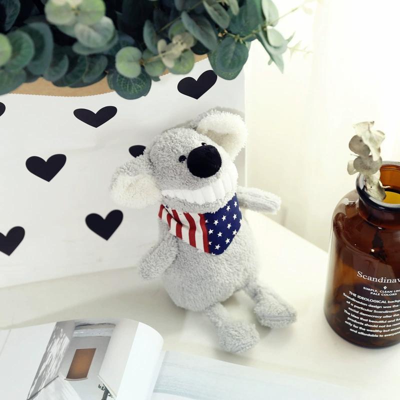 Grey plush toy with a big toothy grin and patriotic scarf, positioned by a vase and leafy plant