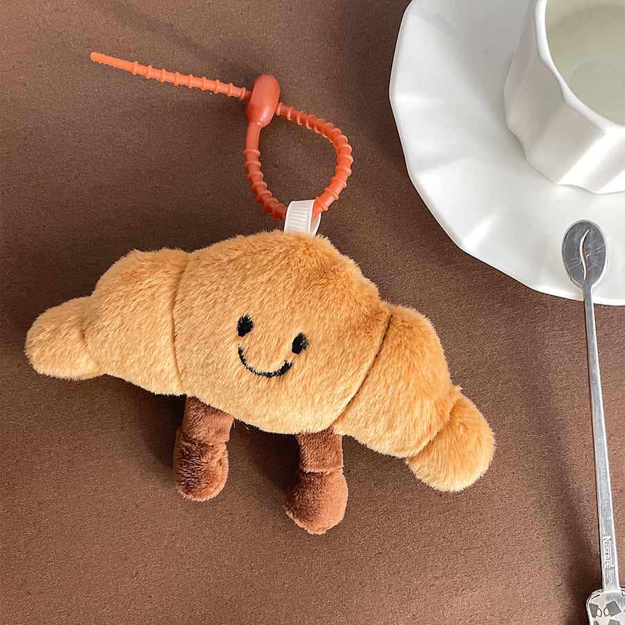 Cute croissant-shaped plush keychain with a smiling face, placed beside a white cup