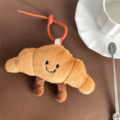Cute croissant-shaped plush keychain with a smiling face, placed beside a white cup