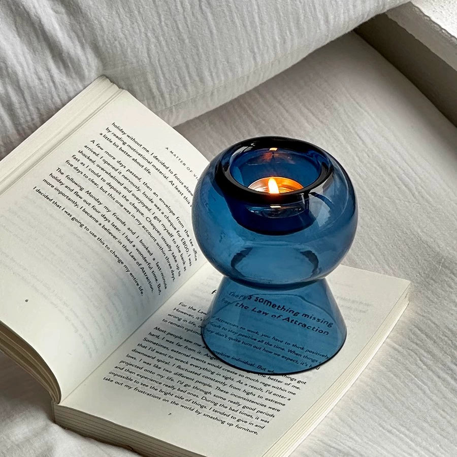 Modern blue glass candle holder with candle burning, placed atop an open book by the bed.