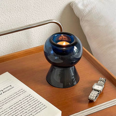 Unique blue glass candle holder glowing with a candle on a wooden nightstand beside a watch and book.