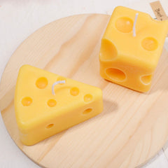 Two cheese-shaped candles, one wedge and one cube style, on a wooden plate, highlighting playful candle design.