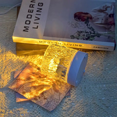 Water ripple aurora lamp lying beside modern living books, softly radiating golden light over textured surfaces and photos.